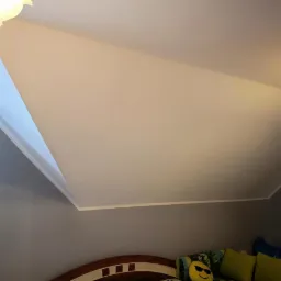 Poddasze po remoncie: skośny sufit pomalowany na biało, ściany szare, widoczny fragment lampy i łóżka z dekoracyjnymi poduszkami.