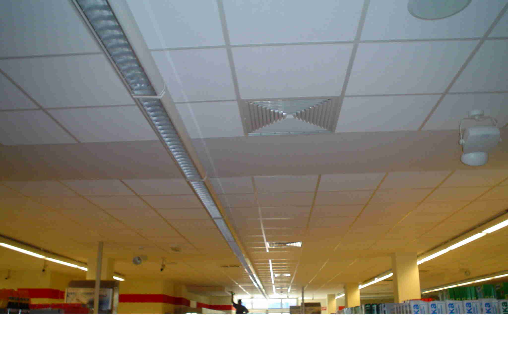 Sufit podwieszany z kratką wentylacyjną w kształcie piramidy i liniowymi lampami LED w przestrzeni handlowej.