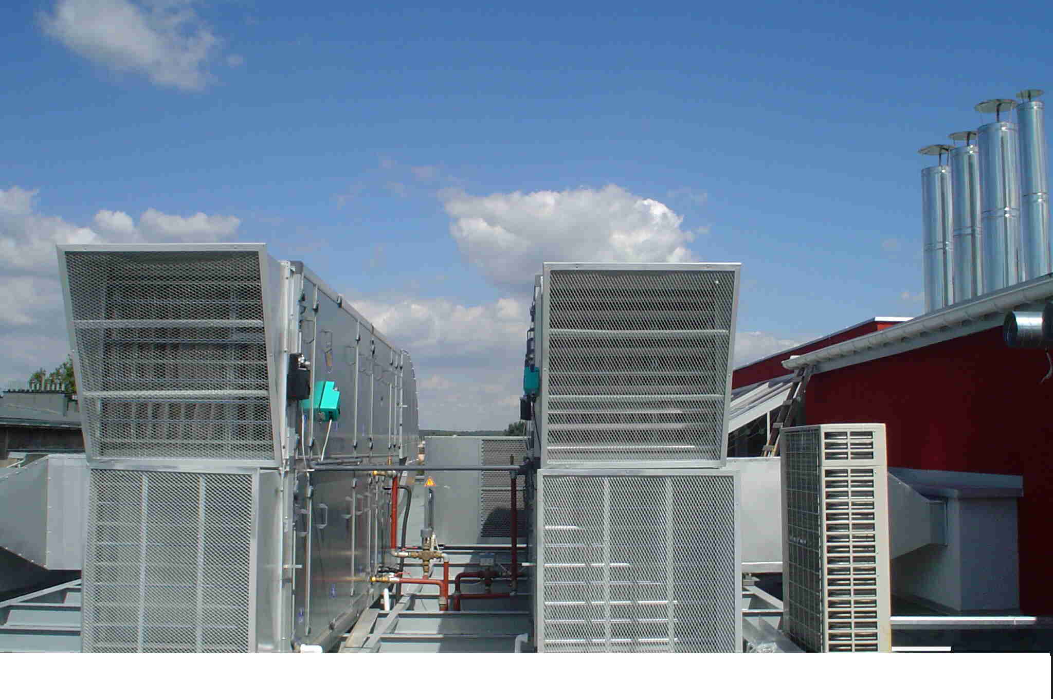 Kompleksowy system klimatyzacji i wentylacji na dachu budynku, widoczne kraty wentylacyjne, rury i elementy automatyki, na tle błękitnego nieba z chmurami.