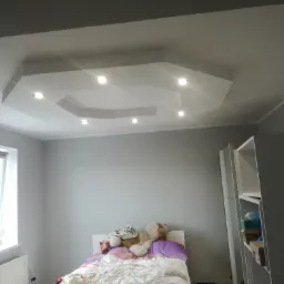 Pokój po malowaniu ścian na szaro, z ozdobnym sufitem podwieszanym z oświetleniem punktowym. Na pierwszym planie łóżko z pluszowym misiem.