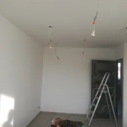 Wnętrze w Dzierżoniowie w trakcie remontu: świeżo ułożone panele podłogowe, pomalowane na biało ściany, wiszące kable oświetleniowe i drabina oparta o zabezpieczone folią drzwi.