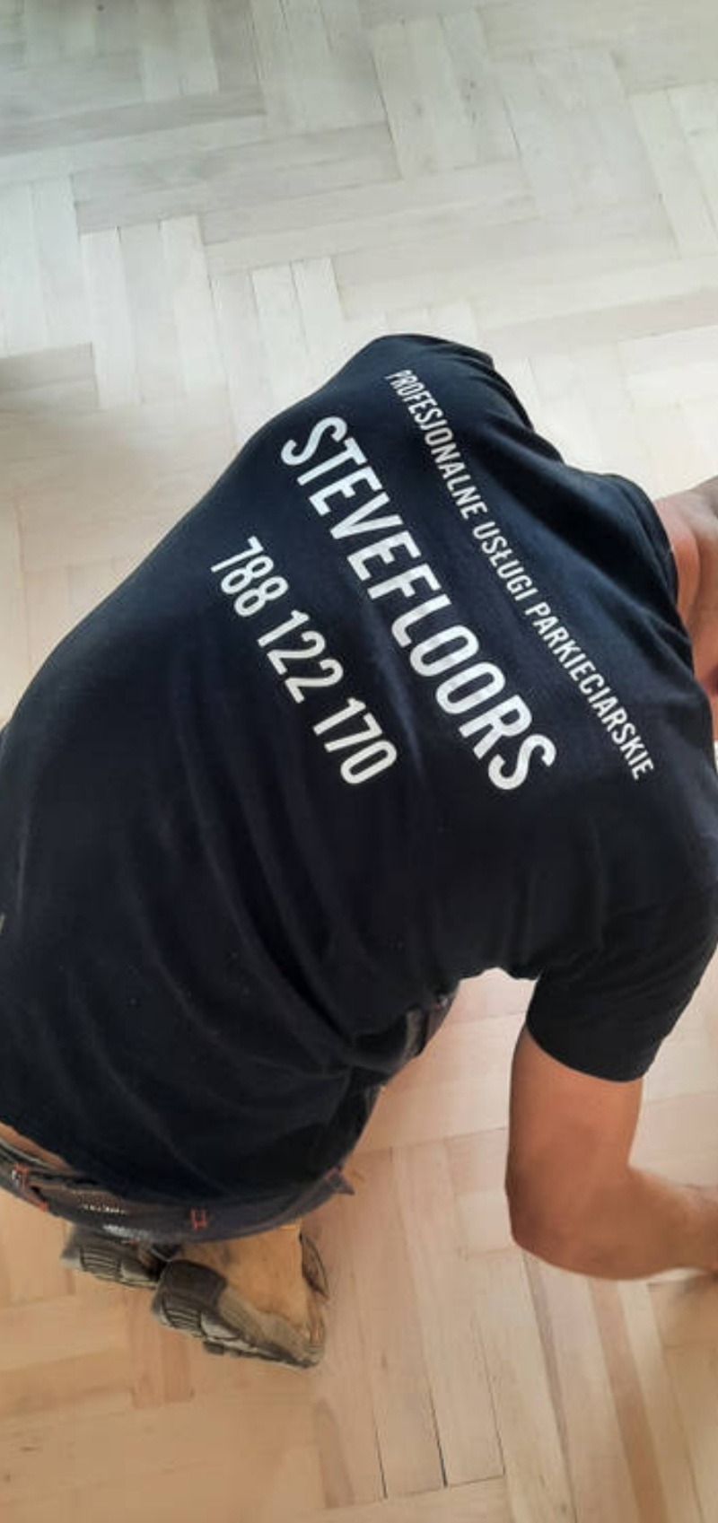 Pracownik w koszulce z logo firmy parkieciarskiej pochylony nad jasną, odnowioną podłogą ułożoną w jodełkę.