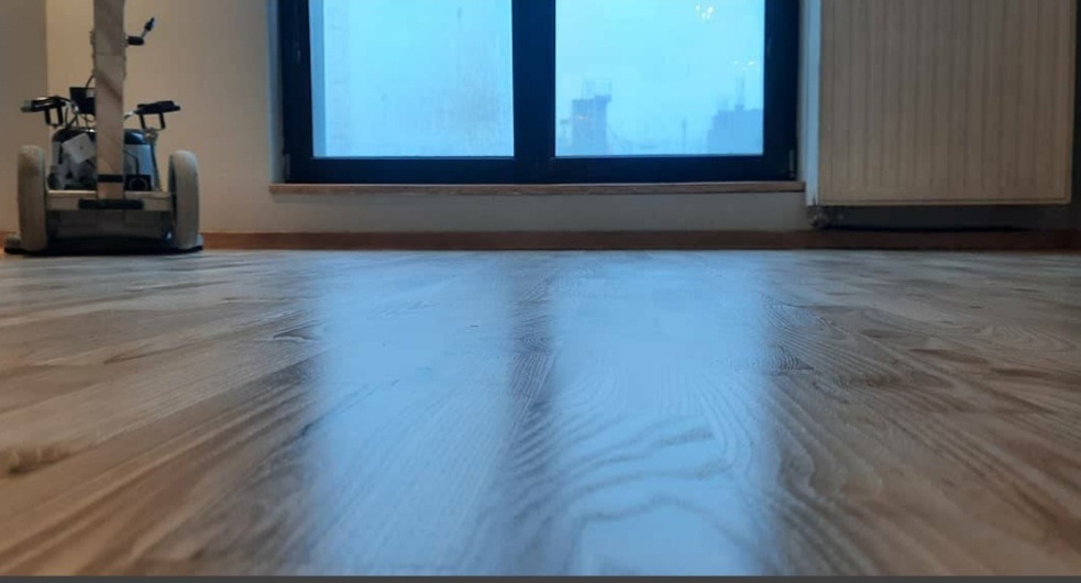Parkiet po cyklinowaniu, widoczna maszyna do cyklinowania w tle, duże okno z widokiem na miasto.