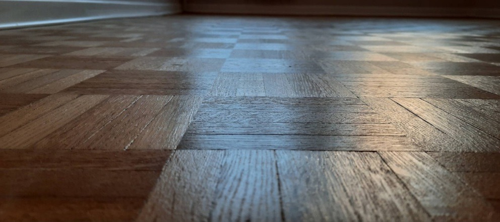 Błyszcząca, odnowiona parkietowa podłoga ułożona w kwadratowy wzór, z widocznymi słojami drewna i delikatnym odbiciem światła.