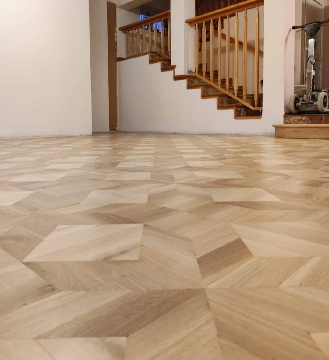 Parkiet ułożony we wzór geometryczny, po renowacji, z widocznymi schodami z drewnianą balustradą w tle i maszyną do cyklinowania.