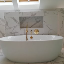 AIRLESS - Nowoczesna, wolnostojąca wanna w łazience wykończonej białym marmurem z złotą armaturą, oświetlona światłem z okna dachowego.