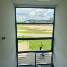 AIRLESS - Widok z klatki schodowej przez duże okno na teren zewnętrzny z budynkami i zielenią, kobieta z koszem przechodzi po chodniku na zewnątrz.