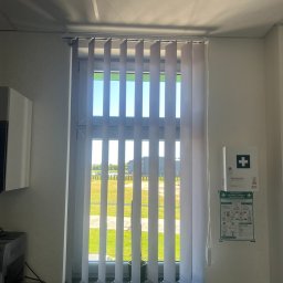 AIRLESS - Wnętrze biura z pionowymi żaluzjami w oknie, widok na zewnątrz z zielonym trawnikiem, grzejnik pod oknem, apteczka pierwszej pomocy na ścianie.