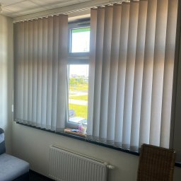 AIRLESS - Zamknięte pionowe żaluzje w jasnym odcieniu szarości, zamontowane w oknie z widokiem na zewnątrz, z elementami dekoracyjnymi na parapecie i grzejnikiem pod oknem.