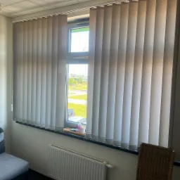 Zamknięte pionowe żaluzje w jasnym odcieniu szarości, zamontowane w oknie z widokiem na zewnątrz, z elementami dekoracyjnymi na parapecie i grzejnikiem pod oknem.