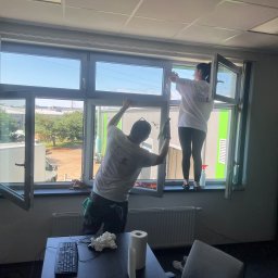 AIRLESS - Dwie osoby w firmowych koszulkach czyszczą okna w biurze, widok na zewnątrz z budynkami przemysłowymi, na biurku klawiatura i rolka papieru.