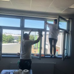 AIRLESS - Dwie osoby w firmowych koszulkach czyszczą duże okno w biurze, używając ścierek i spryskiwacza, widok z wnętrza pomieszczenia na zewnątrz.