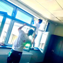 AIRLESS - Dwie osoby w białych koszulkach z logo firmy czyszczą okna w biurze; mężczyzna trzyma ramę okna, a kobieta myje szybę niebieską szmatką.