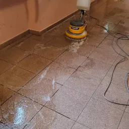 Maszyna do czyszczenia podłóg marki Tenzi w trakcie pracy na brudnych, brązowych płytkach podłogowych, z widoczną pianą i mokrymi plamami.