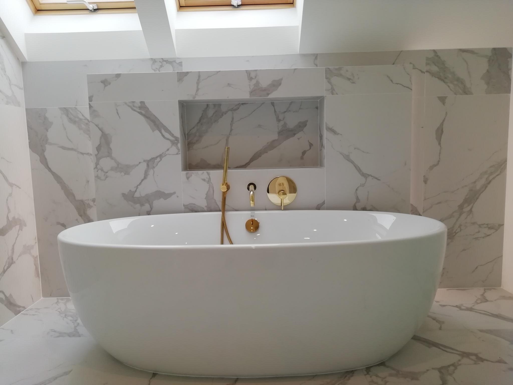Nowoczesna, wolnostojąca wanna w łazience wykończonej białym marmurem z złotą armaturą, oświetlona światłem z okna dachowego.