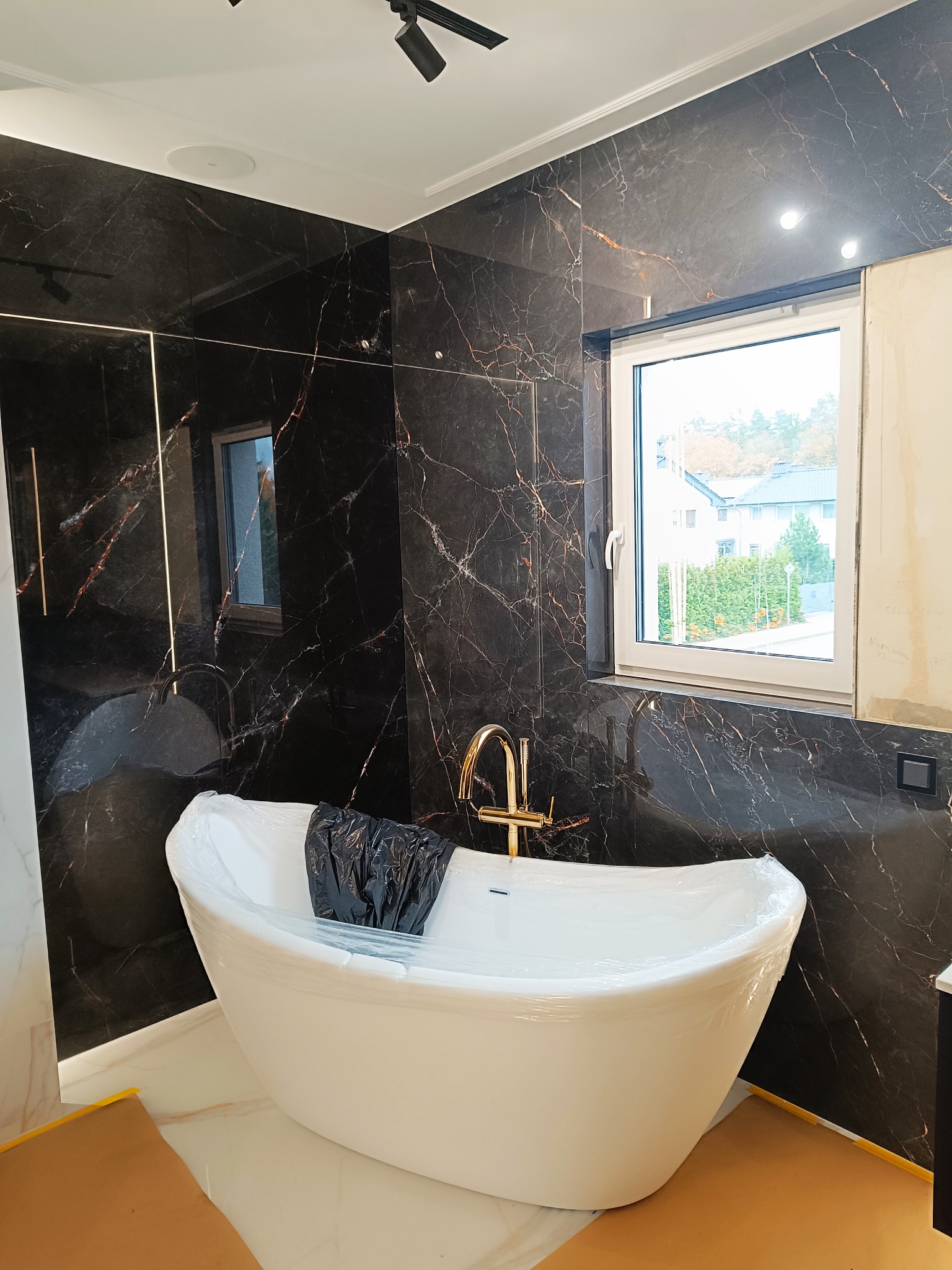 Nowoczesna łazienka z czarnymi marmurowymi płytkami i wolnostojącą białą wanną, jeszcze zabezpieczoną folią ochronną, złota bateria, okno z widokiem na zieleń.