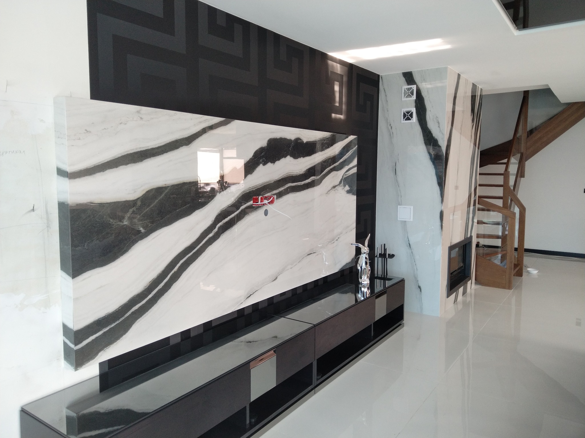 Nowoczesny salon z marmurową ścianą, czarną konsolą pod telewizorem i drewnianymi schodami w tle, wykończony z dbałością o detale.