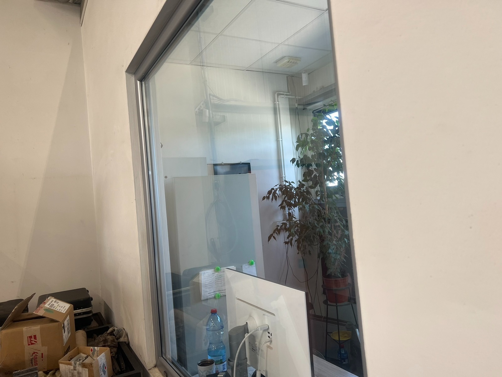 Przeszklona ścianka działowa w biurze, widok z zewnątrz, z widocznym wnętrzem biura: biurko z monitorem, roślina doniczkowa i kartony na podłodze.