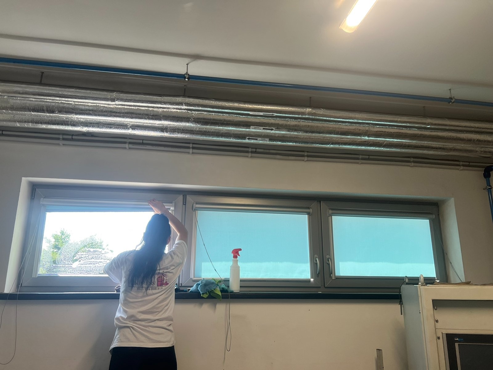 Kobieta w białej koszulce czyści okno w pomieszczeniu biurowym, widoczne rury pod sufitem i butelka ze środkiem czyszczącym na parapecie.