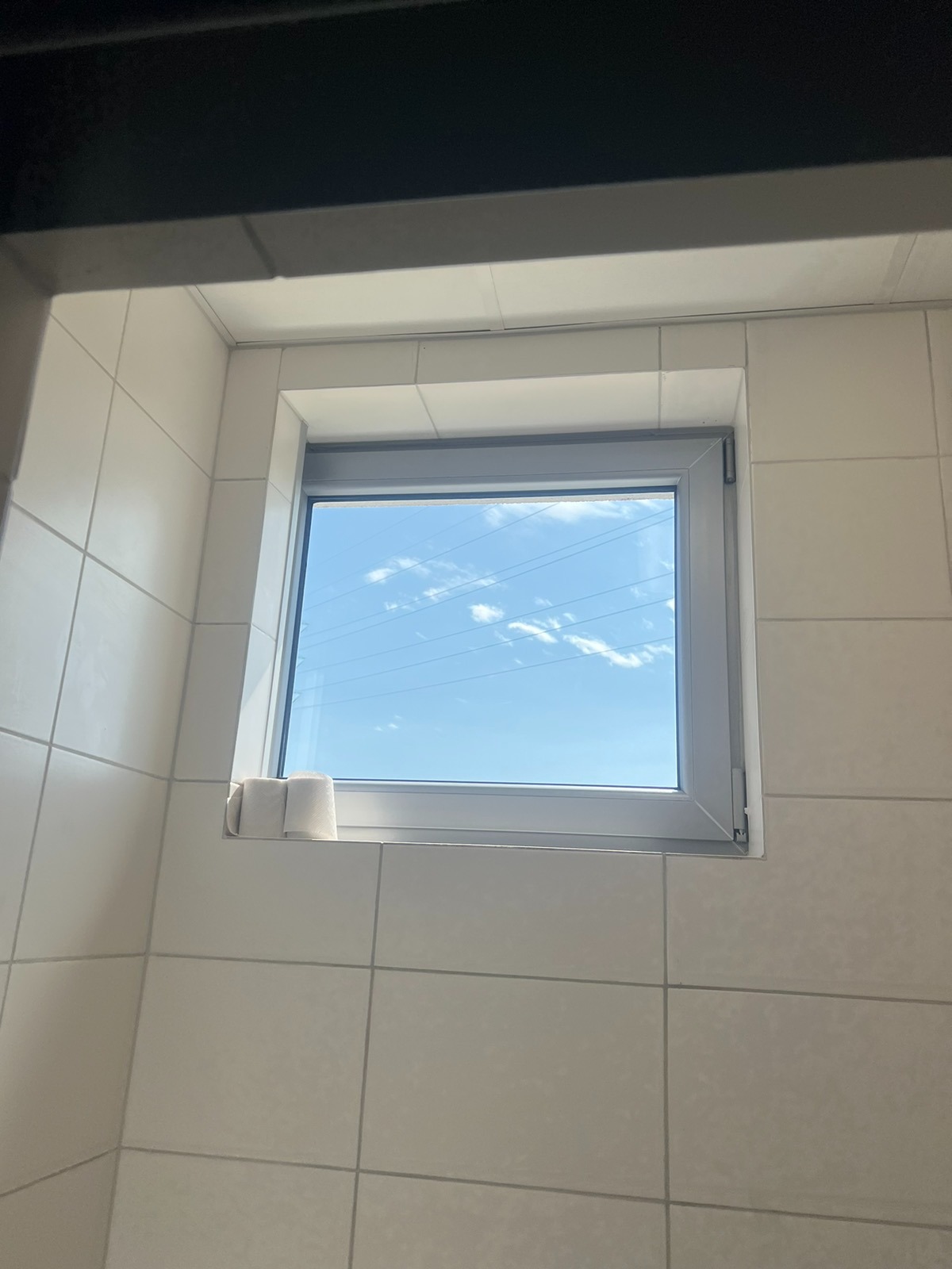 Okienko w łazience wykończonej jasnymi, kwadratowymi płytkami; na parapecie dwie rolki papieru toaletowego, za oknem błękitne niebo z chmurami.