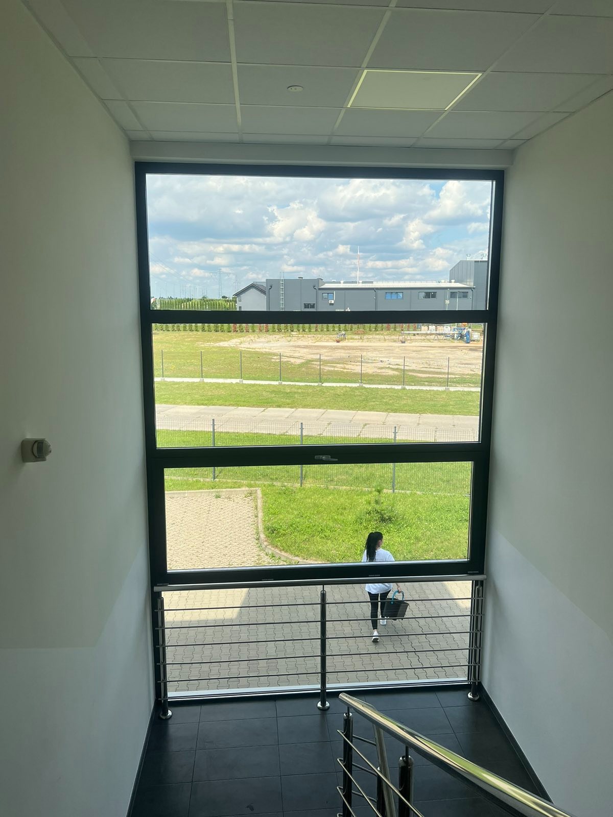 Widok z klatki schodowej przez duże okno na teren zewnętrzny z budynkami i zielenią, kobieta z koszem przechodzi po chodniku na zewnątrz.