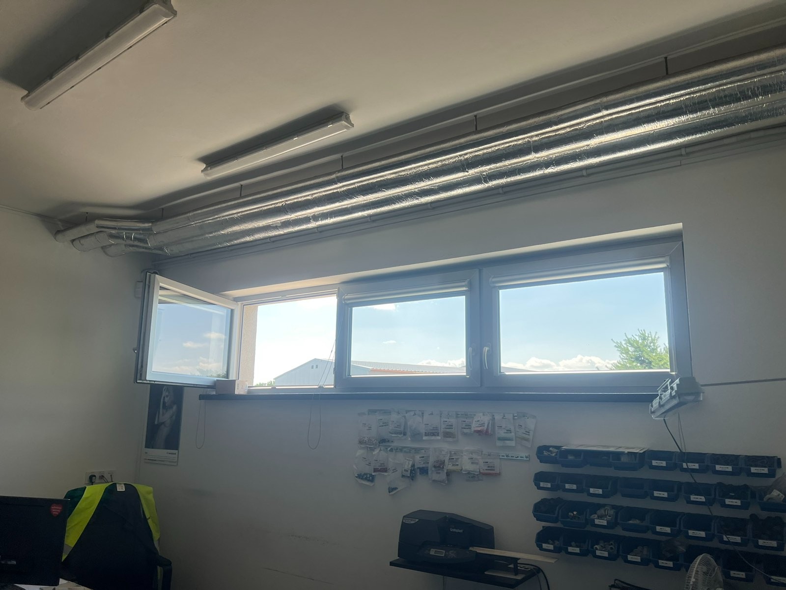 Wnętrze pomieszczenia z oknami, widoczne rury wentylacyjne pod sufitem, otwarte okno po lewej stronie, na ścianie półki z pojemnikami i akcesoriami.