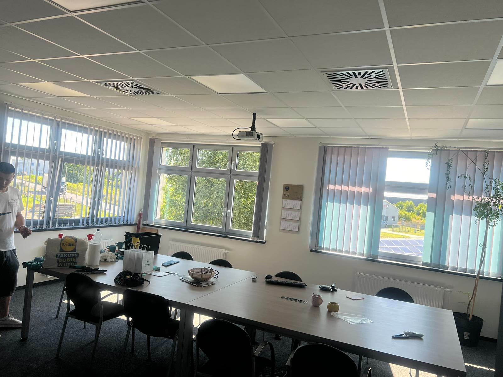 Jasne biuro z dużym stołem konferencyjnym, roletami w oknach i widocznymi śladami porządkowania po sprzątaniu, w tle torba z logo sklepu.