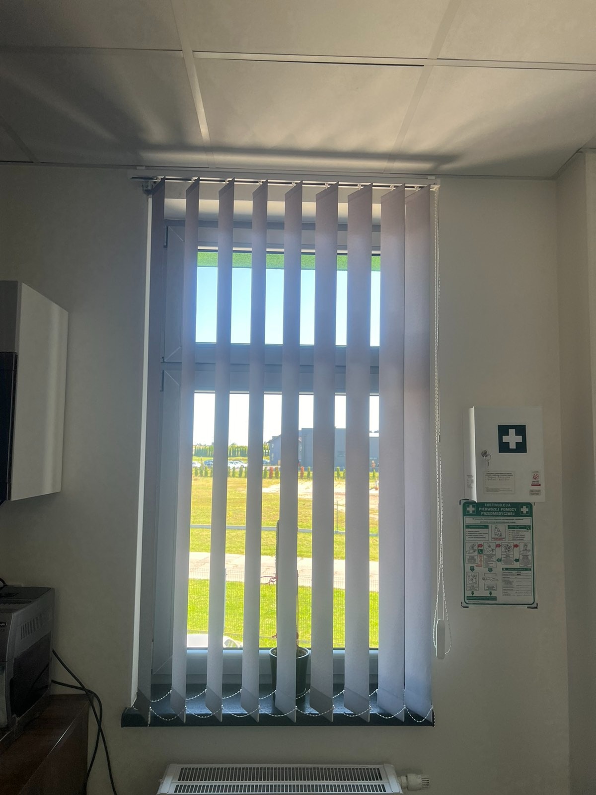 Wnętrze biura z pionowymi żaluzjami w oknie, widok na zewnątrz z zielonym trawnikiem, grzejnik pod oknem, apteczka pierwszej pomocy na ścianie.