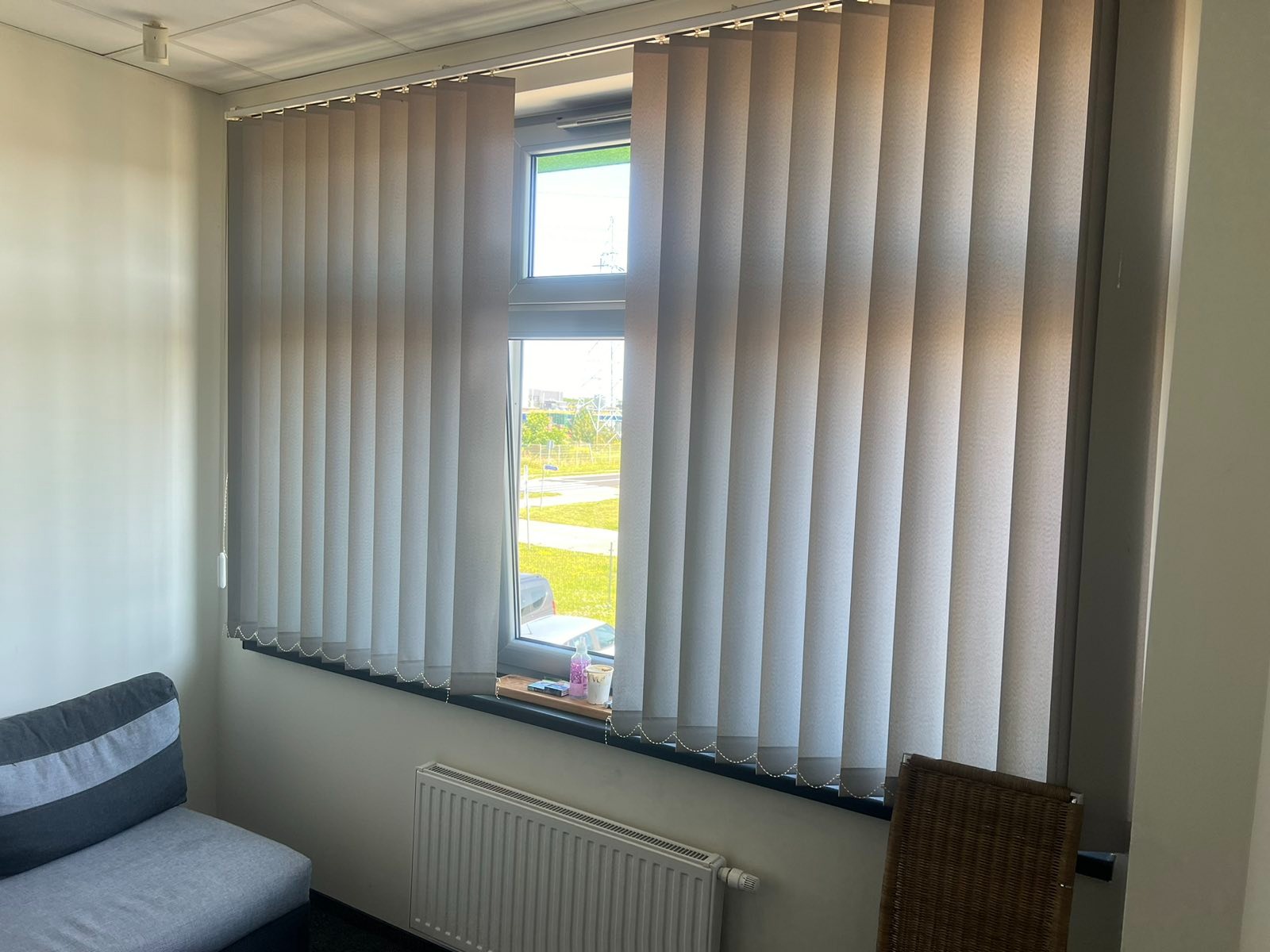 Zamknięte pionowe żaluzje w jasnym odcieniu szarości, zamontowane w oknie z widokiem na zewnątrz, z elementami dekoracyjnymi na parapecie i grzejnikiem pod oknem.