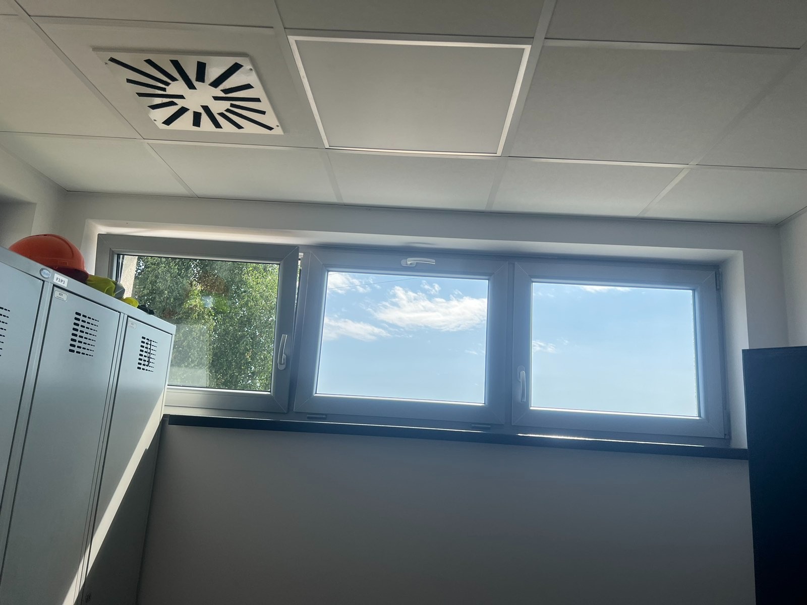 Wnętrze biura z oknem, widokiem na zewnątrz i fragmentem szafki pracowniczej z pomarańczowym kaskiem.