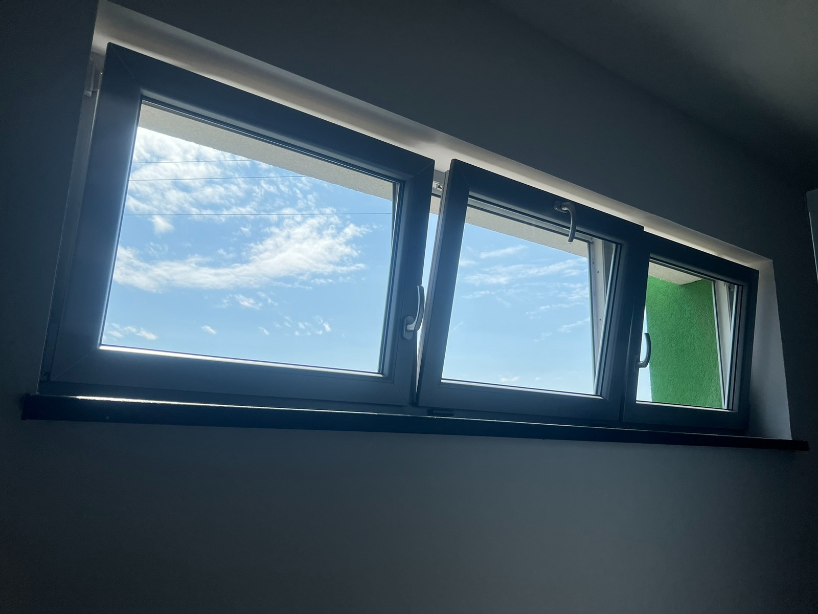 Trzyczęściowe okno PCV z szarymi ramami, dwa skrzydła uchylone, widok na błękitne niebo z chmurami, zielona ściana widoczna przez jedno z okien.