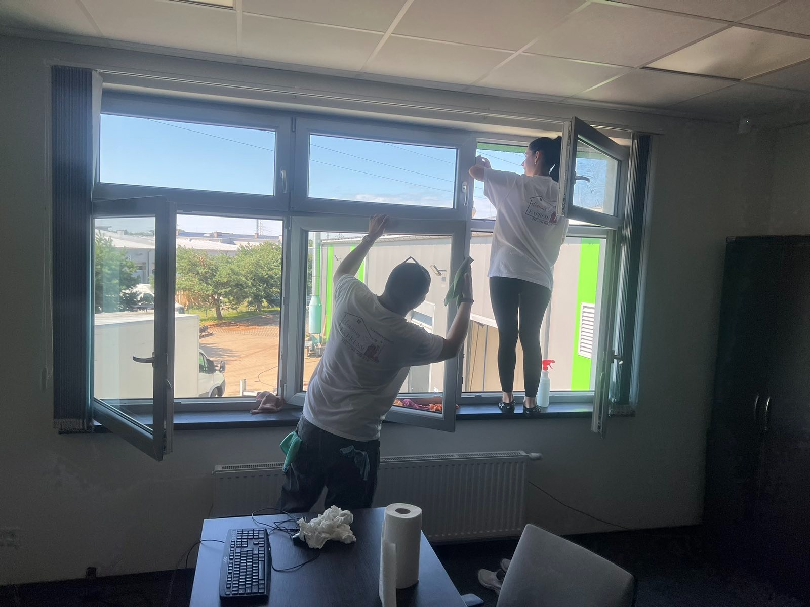 Dwie osoby w firmowych koszulkach czyszczą okna w biurze, widok na zewnątrz z budynkami przemysłowymi, na biurku klawiatura i rolka papieru.