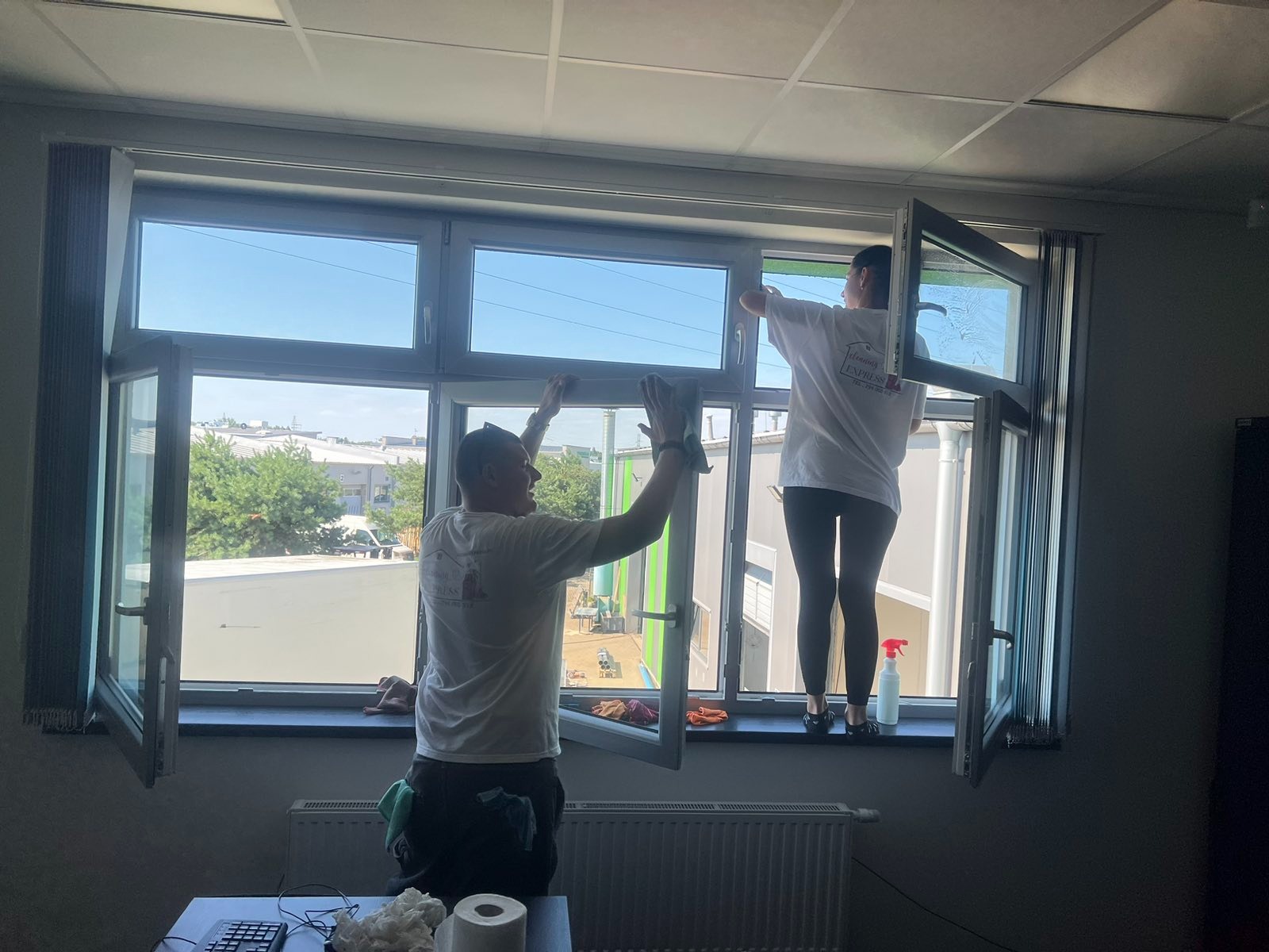 Dwie osoby w firmowych koszulkach czyszczą duże okno w biurze, używając ścierek i spryskiwacza, widok z wnętrza pomieszczenia na zewnątrz.