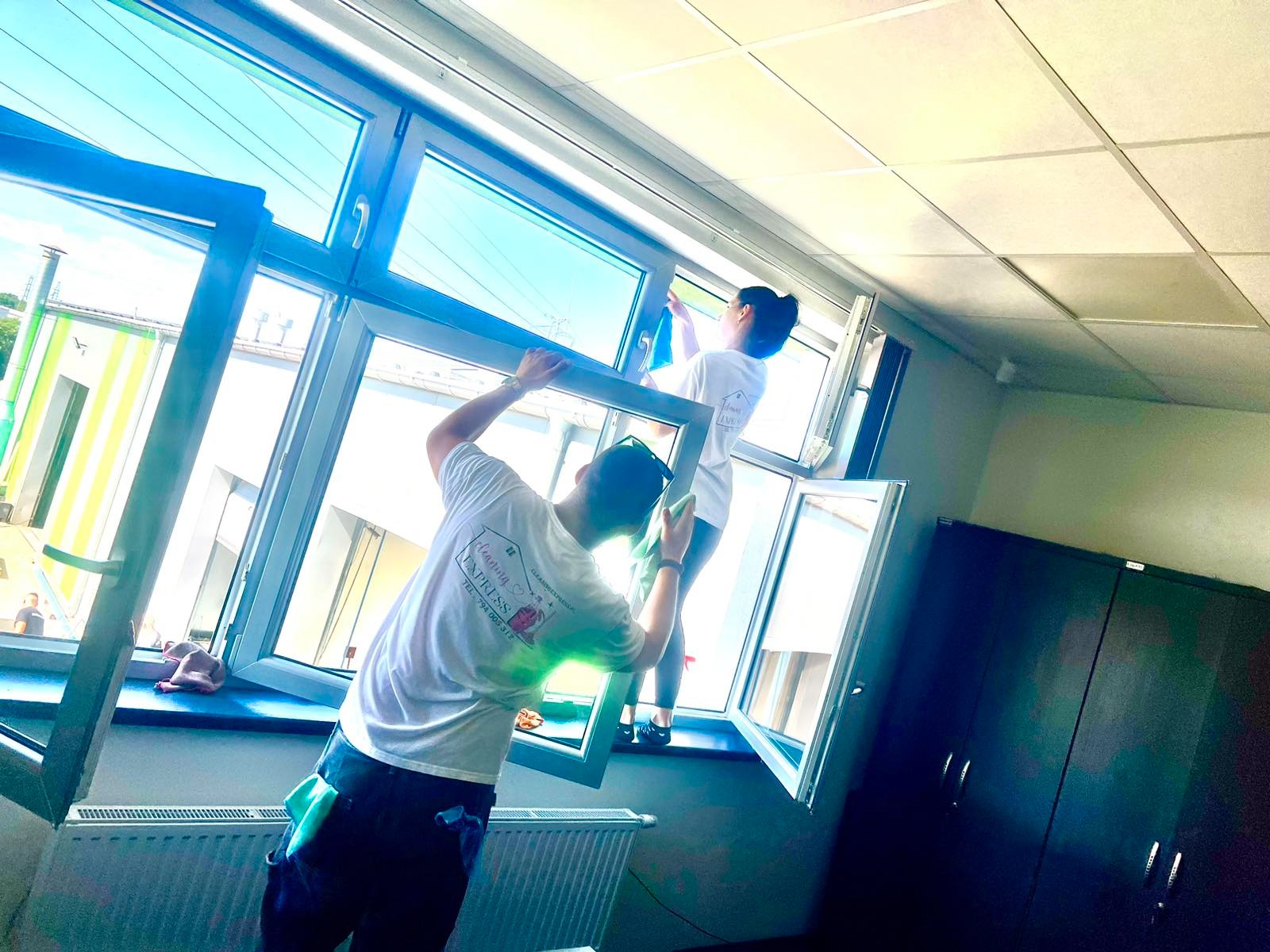 Dwie osoby w białych koszulkach z logo firmy czyszczą okna w biurze; mężczyzna trzyma ramę okna, a kobieta myje szybę niebieską szmatką.