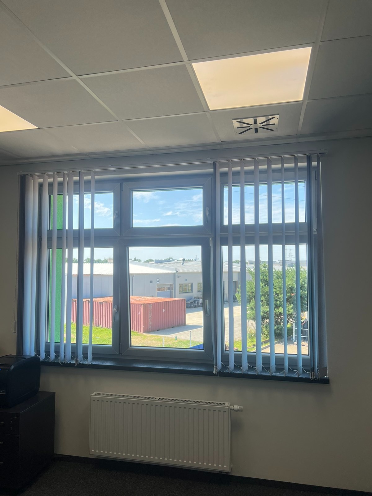 Wnętrze biura w Szczecinie z oknem z żaluzjami pionowymi, widok na kontenery magazynowe i budynek przemysłowy, grzejnik pod oknem.