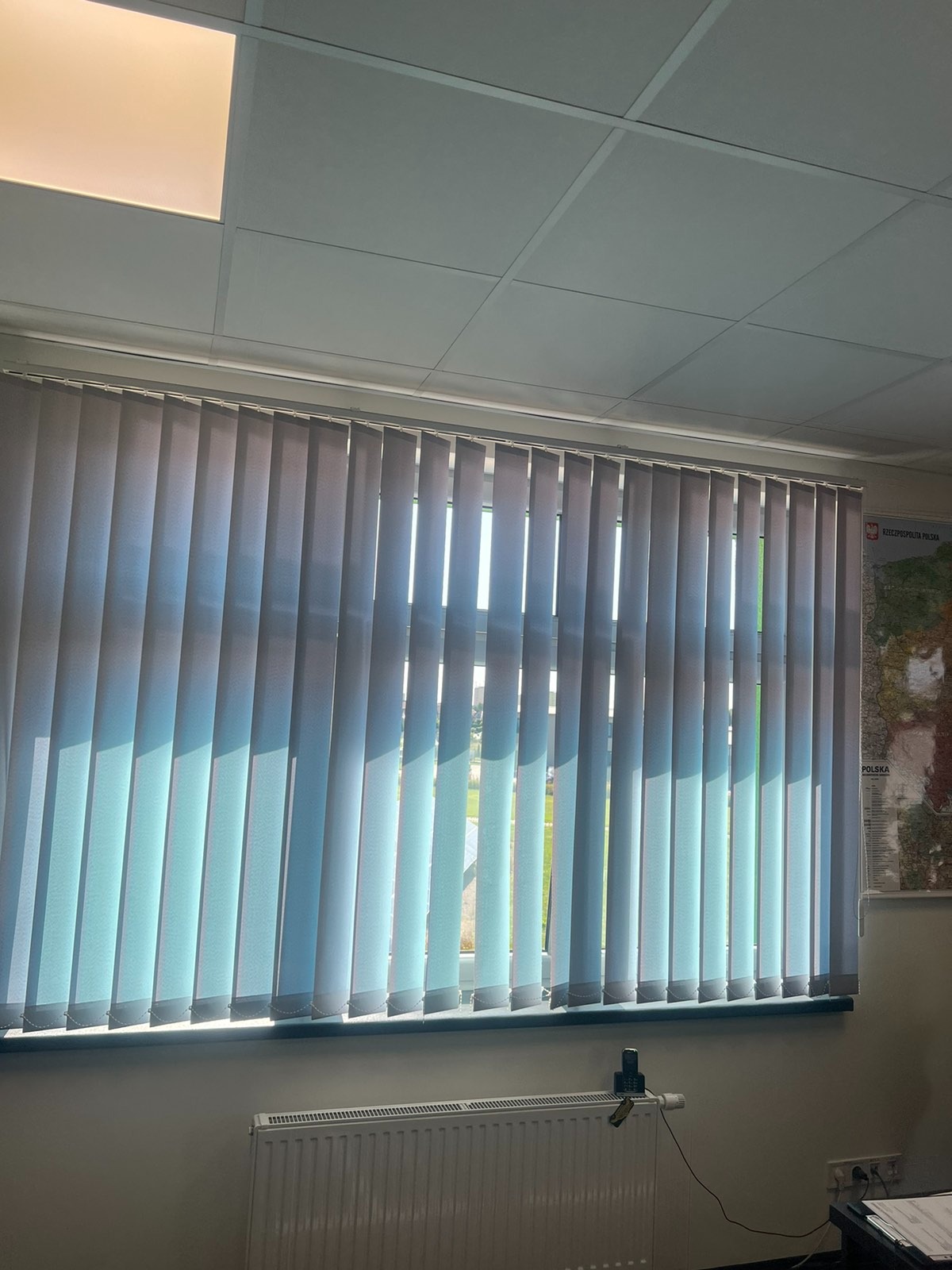 Białe pionowe żaluzje w biurze, częściowo zasłonięte, wpadające światło słoneczne, widoczny grzejnik i mapa Polski na ścianie.