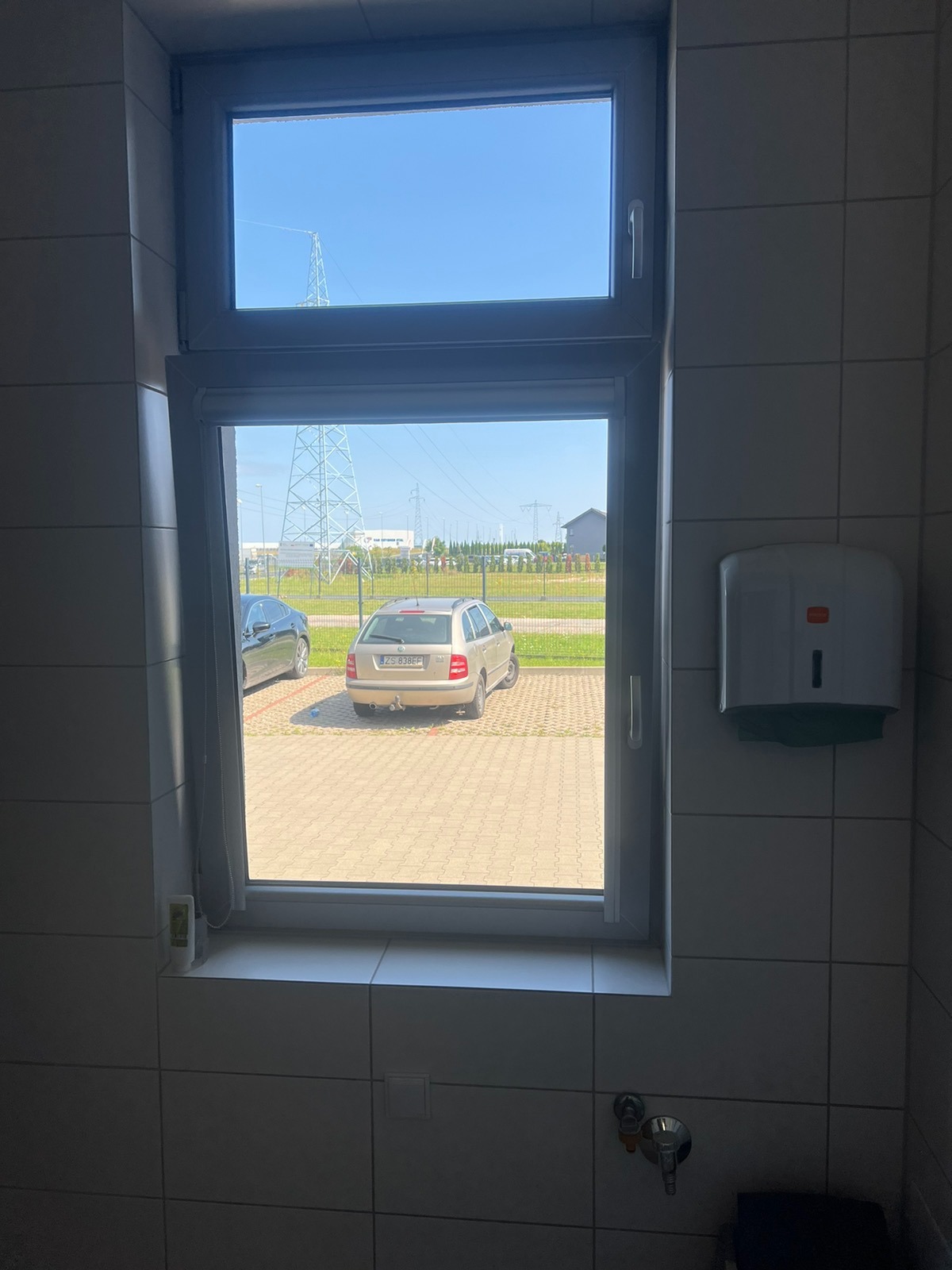 Widok z okna łazienki na parking z dwoma samochodami i słupem wysokiego napięcia w tle, widoczny dozownik na ręczniki na kafelkowanej ścianie.