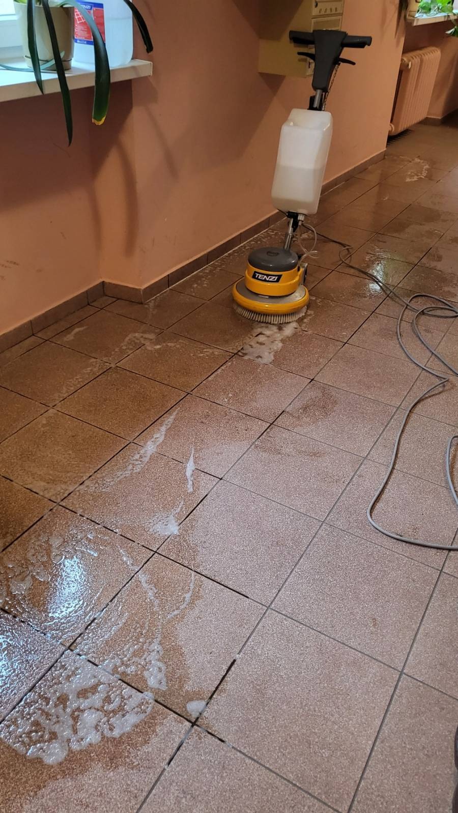 Maszyna do czyszczenia podłóg marki Tenzi w trakcie pracy na brudnych, brązowych płytkach podłogowych, z widoczną pianą i mokrymi plamami.