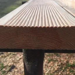 Przekrój deski tarasowej z ryflowaną powierzchnią, widoczne słoje drewna i profil krawędzi, umieszczonej na metalowym wsporniku.