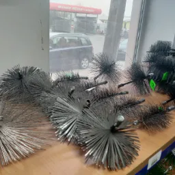 Szczotki kominiarskie z metalowym włosiem i uchwytami na drewnianej półce, widok z okna na myjnię bezdotykową.