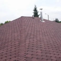 Pracownik w niebieskiej koszulce i czapce, klęczący na dachu pokrytym brązowym gontem bitumicznym, wykonuje prace wykończeniowe przy kominie wentylacyjnym.