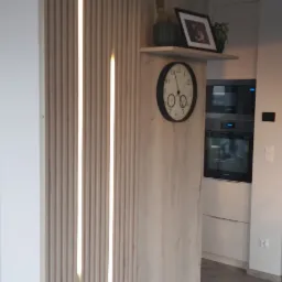 Drewniana ścianka działowa z pionowymi listwami i wbudowanym oświetleniem LED, półka z dekoracjami (kaktus, ramka ze zdjęciem, roślina), zegar na ścianie, w tle widoczna zabudowa kuchenna w jasnych...