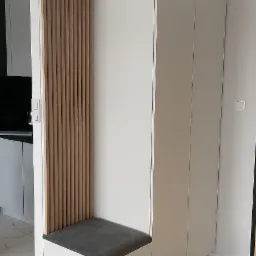 Biała szafa wnękowa z siedziskiem i drewnianymi lamelami, szara tapicerka siedziska, marmurowe płytki podłogowe.