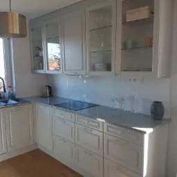 Jasna kuchnia z szarym blatem, przeszklonymi szafkami górnymi i złotymi uchwytami, oświetlona naturalnym światłem padającym z okna.