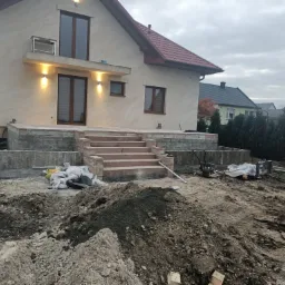 Nowoczesny dom jednorodzinny z beżową elewacją i oświetleniem zewnętrznym, w trakcie budowy tarasu z kamiennymi schodami; widoczne maszyny budowlane i hałda ziemi.