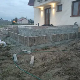 Wznoszenie fundamentów pod taras przy domu jednorodzinnym z wykorzystaniem betonowych bloczków i kamiennej kostki, widoczny sprzęt budowlany i teren budowy.