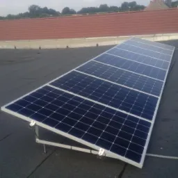 Panele fotowoltaiczne zamontowane na płaskim dachu z widocznymi elementami mocującymi i kablem, w tle ceglany dach innego budynku.