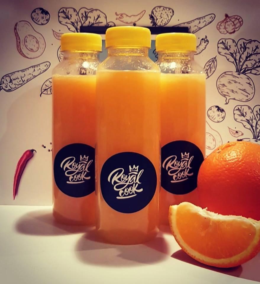 Trzy butelki świeżo wyciskanego soku pomarańczowego z logo 'Royal Cook', podane z całą pomarańczą i jej plasterkiem, na tle z rysunkami warzyw i owoców. Obok butelek leży papryczka chili.