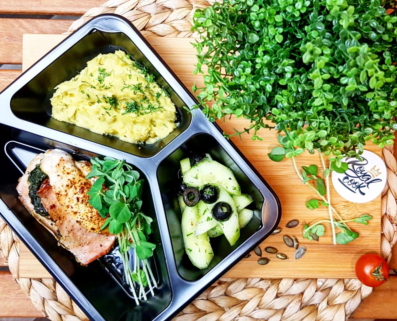 Kompozycja posiłku w czarnym pojemniku z trzema przegródkami: puree z zielonymi ziołami, filet z kurczaka z szynką i szpinakiem, sałatka z ogórków z oliwkami, wszystko sfotografowane z góry...