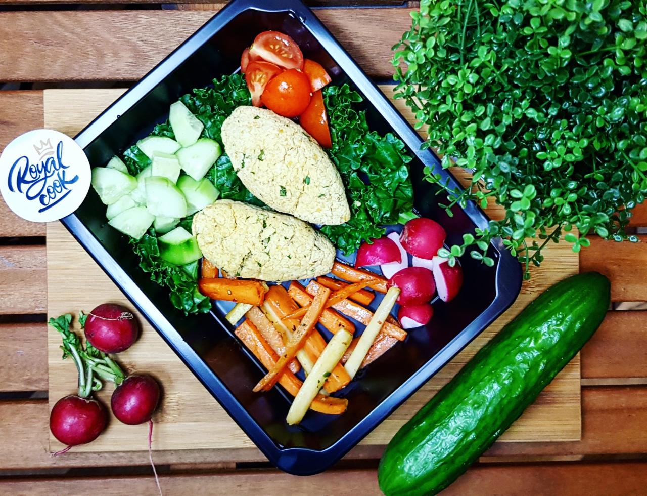 Dietetyczny posiłek w czarnym pojemniku: dwa kotlety, marchewki, rzodkiewki, ogórki, pomidorki koktajlowe i zielenina na drewnianym tle, obok zielony ogórek i roślina doniczkowa, naklejka Royal Cook.